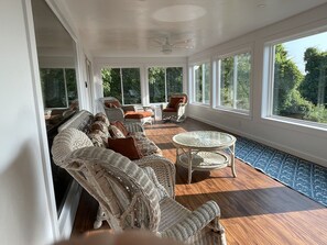 Ein sonnendurchflutetes Zimmer mit Korbmöbeln, großen Fenstern und Blick auf Grün.