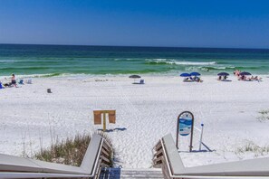 A beach with white sand, ocean waves, and beach chairs. There is a sign with safety information.