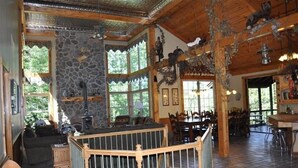 Ein rustikales Blockhaus-Interieur mit Steinwänden, Holzbalken und einem Kamin aus Stein. Es gibt einen Essbereich mit Holztischen und -stühlen sowie eine Bar mit Hockern.