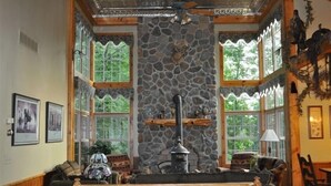 Ein gemütliches Interieur mit Kamin aus Stein, Holzbalken und großen Fenstern mit Blick ins Grüne.