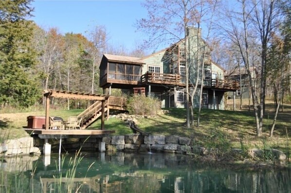 A rustic cabin with a wooden deck and a pond in front.