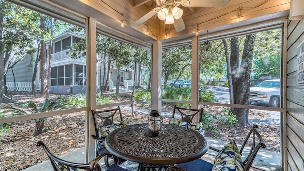 Une terrasse couverte avec une table et des chaises rondes en métal, de grandes fenêtres offrant une vue sur les environs et un ventilateur de plafond.