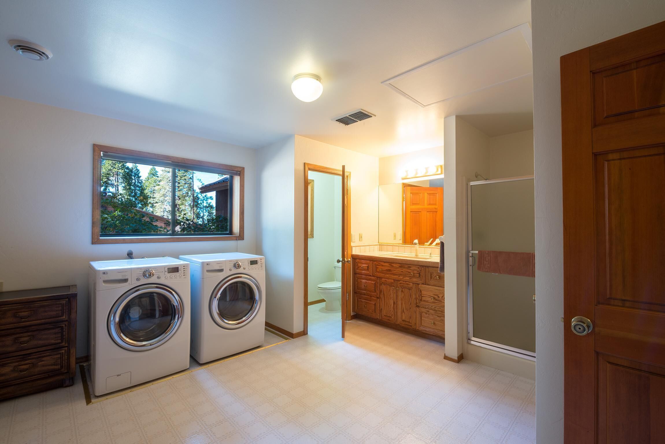 A laundry room with two washing machines, a window with a view of trees, and a wooden cabinet with a mirror.