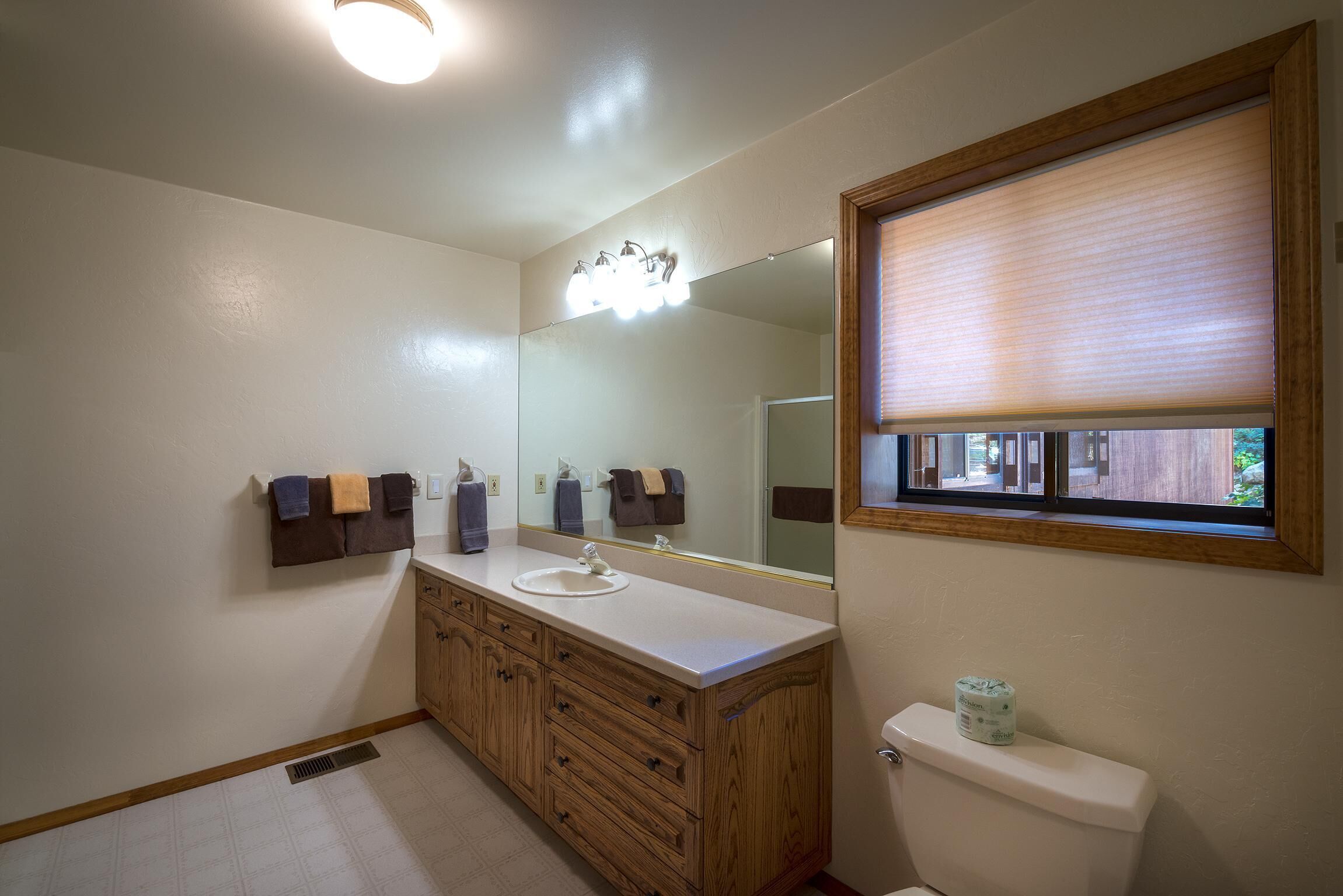 Un baño con tocador de madera, un lavabo blanco, un espejo con iluminación, un inodoro y una ventana con persianas.