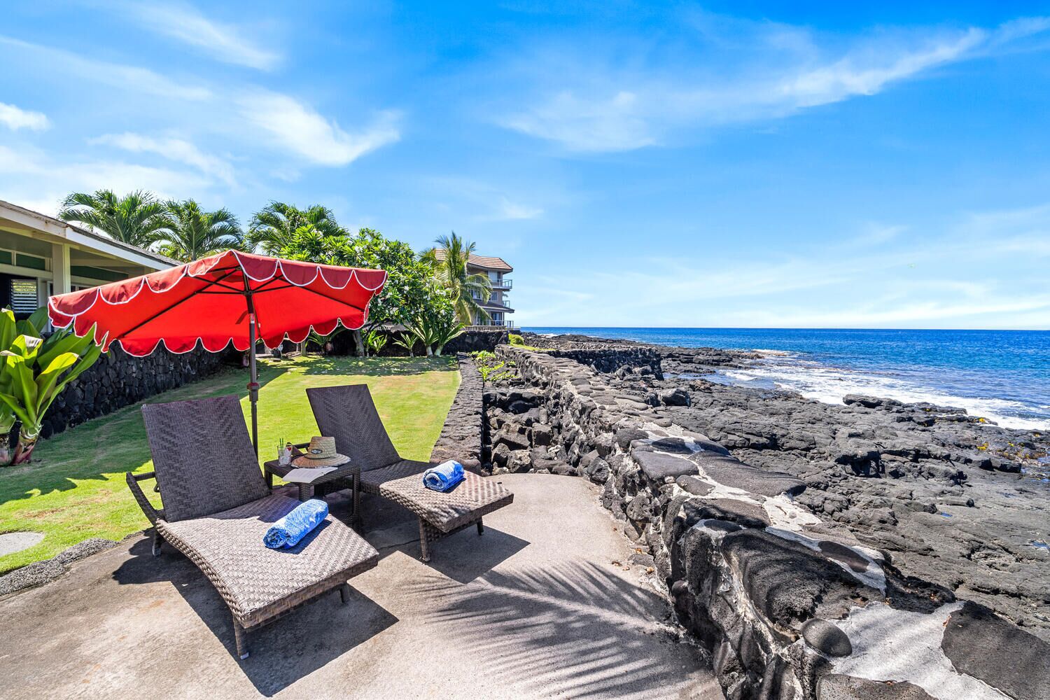 Une terrasse au bord de la plage, dotée de chaises longues et d’un parasol rouge, offrant une vue sur une côte rocheuse et l’océan.