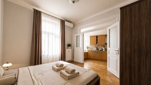 Ein Hotelzimmer mit Bett, Nachttisch, Lampe, einem Fenster mit Vorhängen sowie einer sichtbaren Küchenzeile durch eine geöffnete Tür und einem Holzboden.