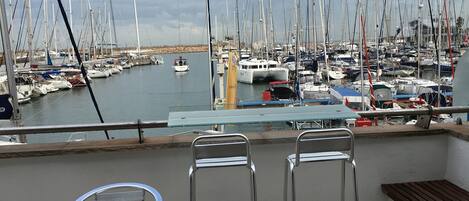 A terrace with a glass table and metal chairs overlooking a marina with numerous sailboats.