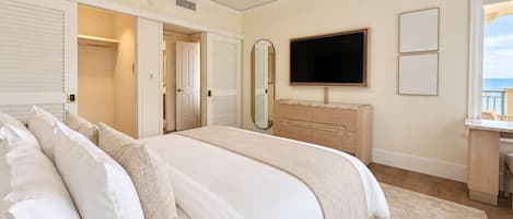 A bedroom with a large bed, a ceiling fan, a TV, a dresser, and a view of the sea.