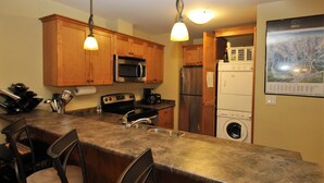 A kitchen with wooden cabinets, a microwave, a refrigerator, a washing machine, and a bar area with chairs. - Spectacular Top Floor 2 Br W/ Hot Tub and Panoramic View (Golden)