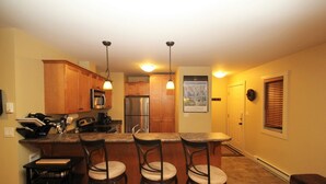 A kitchen with wooden cabinets, a stainless steel refrigerator, a microwave, and a coffee maker. There are three bar stools and a small table. - Spectacular Top Floor 2 Br W/ Hot Tub and Panoramic View (Golden)