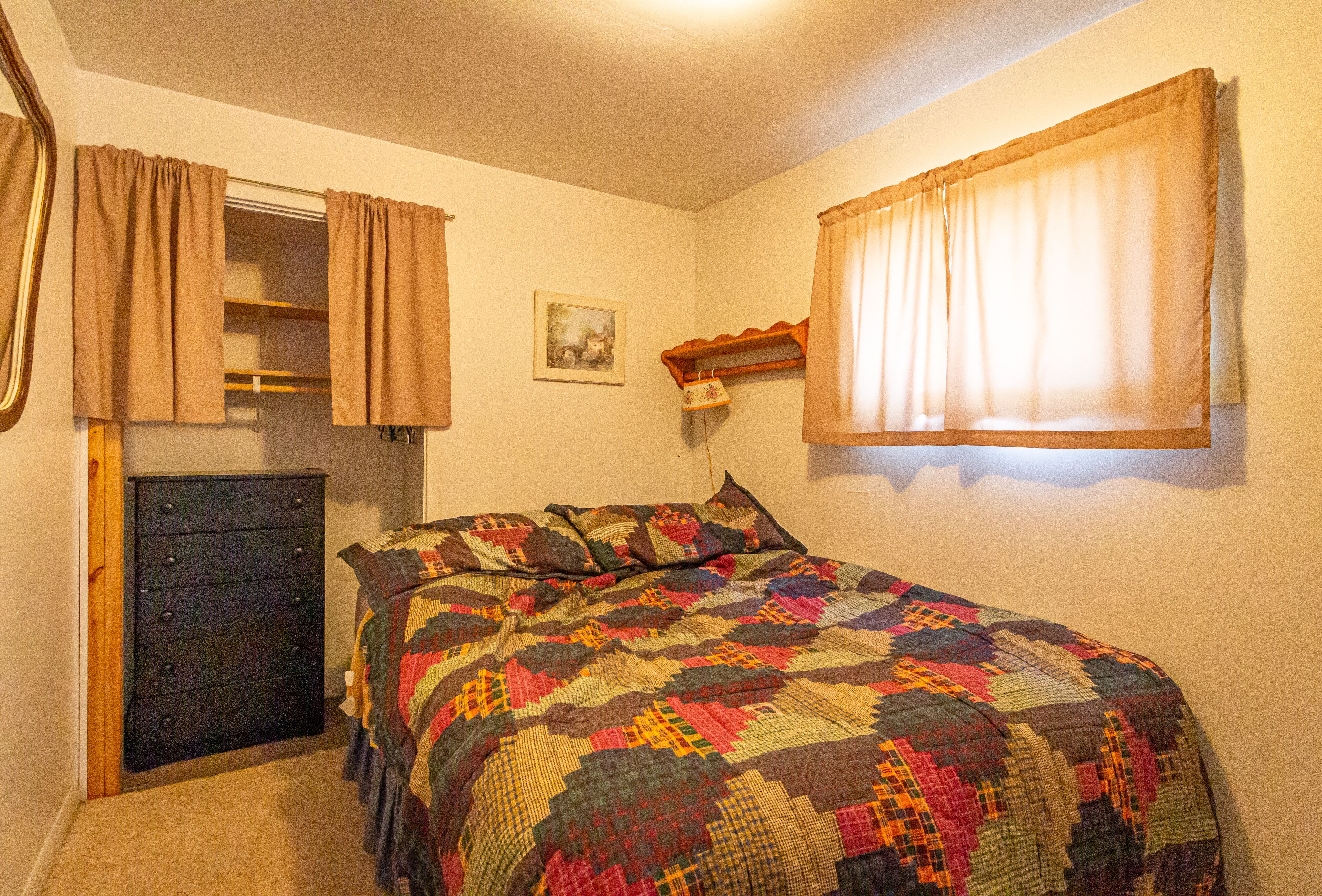 A bedroom with a bed, a dresser, a window with curtains, and a wall-mounted shelf with a lamp.