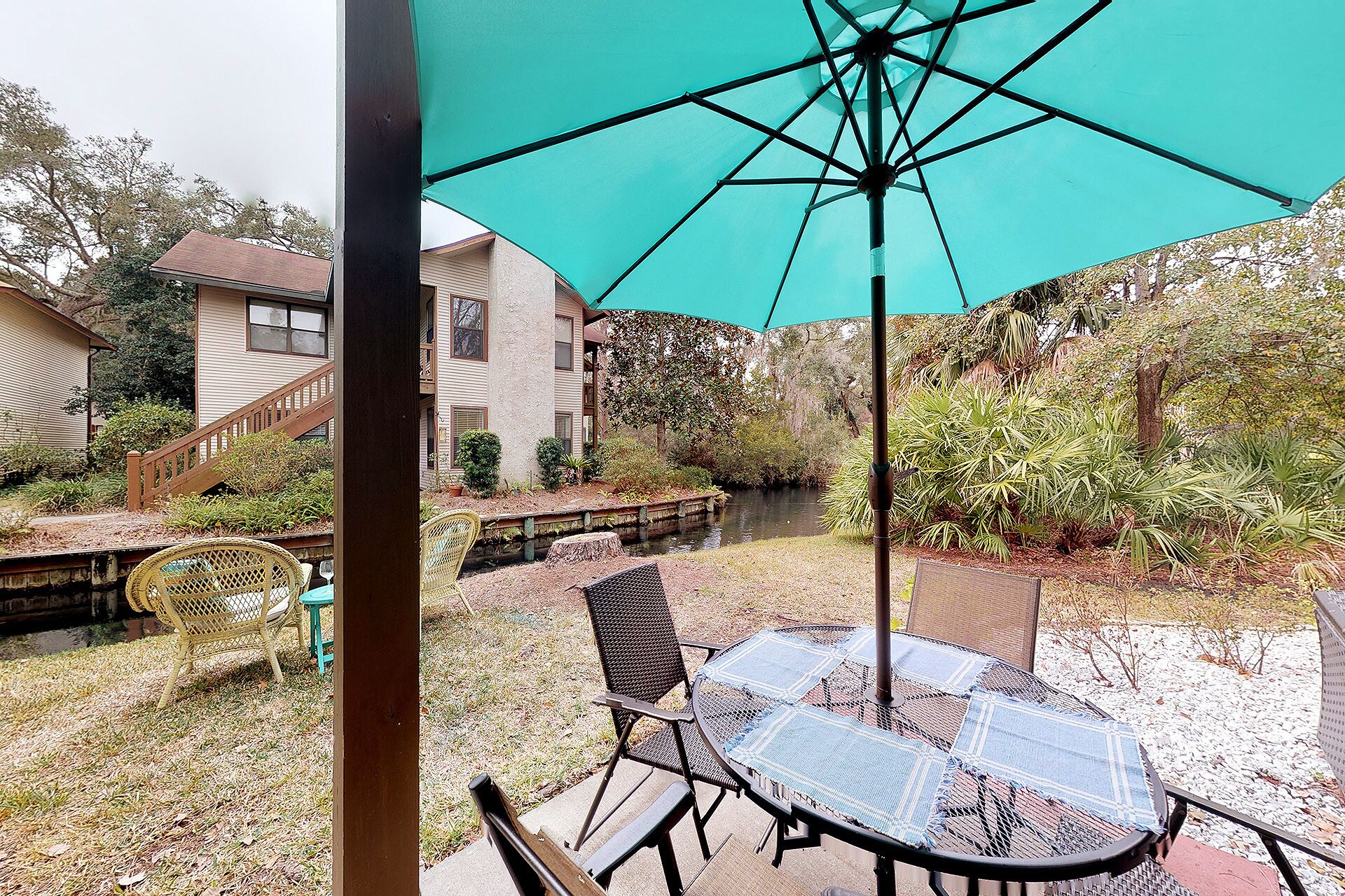 A patio with a table and chairs, a turquoise umbrella, and a view of a house and garden.