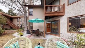 Un bâtiment à deux étages doté d’un balcon, d’un espace extérieur aménagé avec des chaises et une table en osier, et d’un grand parasol offrant de l’ombre.