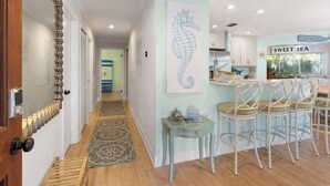 Un couloir avec une porte en bois, un tapis à motifs, un tableau représentant un hippocampe et un coin cuisine avec une pancarte indiquant « THÉ SUCRÉ ».