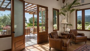 An open-plan living area with a wooden floor, leather furniture, and large windows offering a view of a garden and mountains. - Luxury Napa Valley Estate Surrounded by Vineyards (Calistoga)