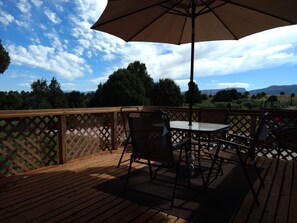 Una terraza de madera con una mesa, sillas, un paraguas y vistas a árboles y montañas.