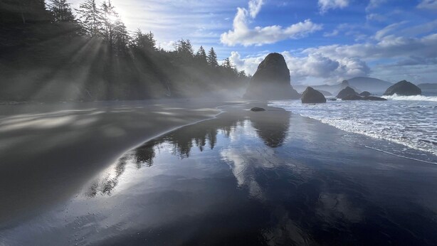 A beach with calm waters reflecting the sky, surrounded by forested hills and large rock formations. - China Mountain House - Ocean Views & Modern Design (Port Orford)