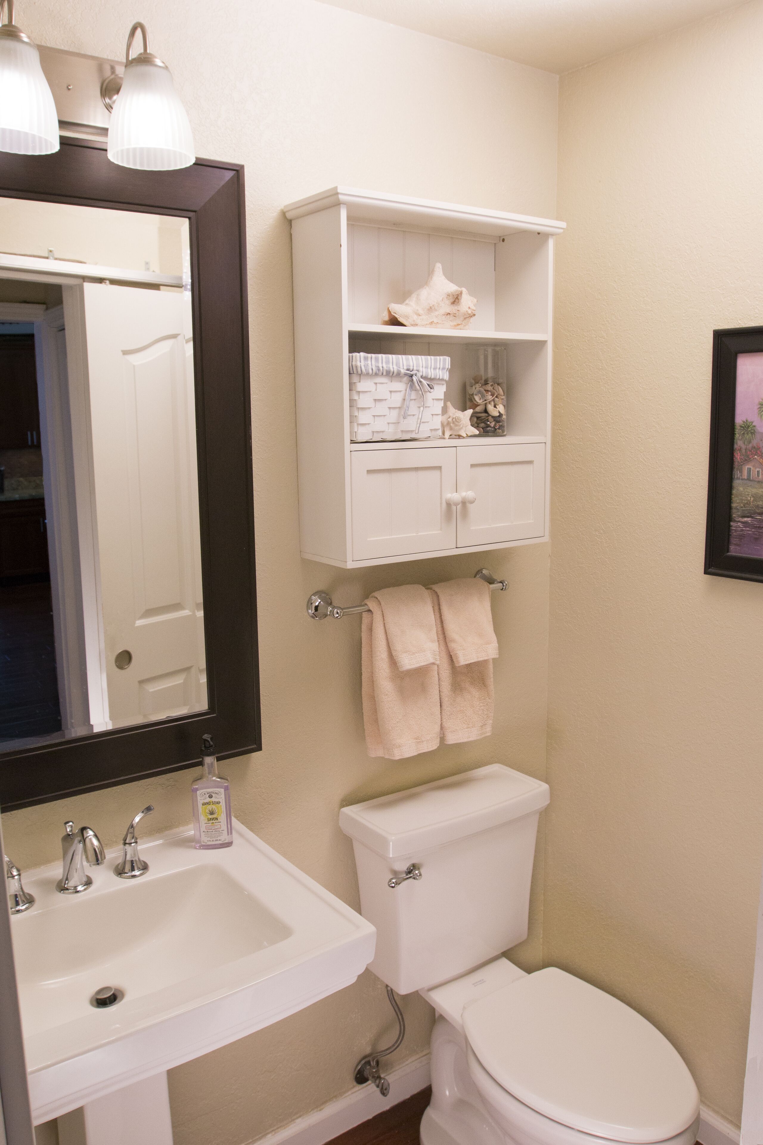 A bathroom with a white sink, toilet, and a mirror above the sink. There are two towels hanging on the wall and a shelf with decorative items.