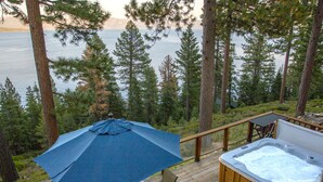 Una terraza con un jacuzzi, una sombrilla y una mesa con sillas, con vistas a un lago rodeado de árboles.
