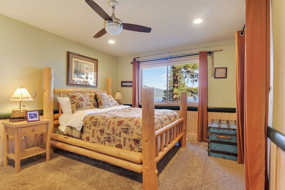 Un dormitorio con cama de madera, ventilador de techo, ventana con cortinas, mesita de noche con lámpara y maleta apoyada en la pared.
