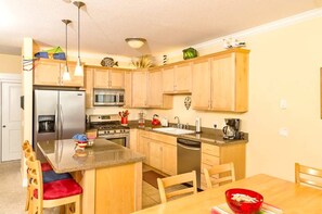 A compact kitchen with wooden cabinets, a stainless steel refrigerator, a microwave, a stove, and a sink. There is a dining area with a wooden table and chairs. - Pacific Life Condo, Ocean View Hot Tub & Pool (Lincoln City)