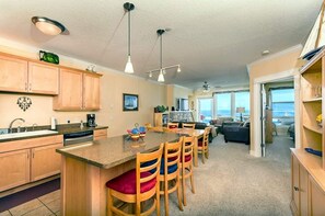 A compact kitchen with wooden cabinets, a central island with bar stools, and a dining area. - Pacific Life Condo, Ocean View Hot Tub & Pool (Lincoln City)