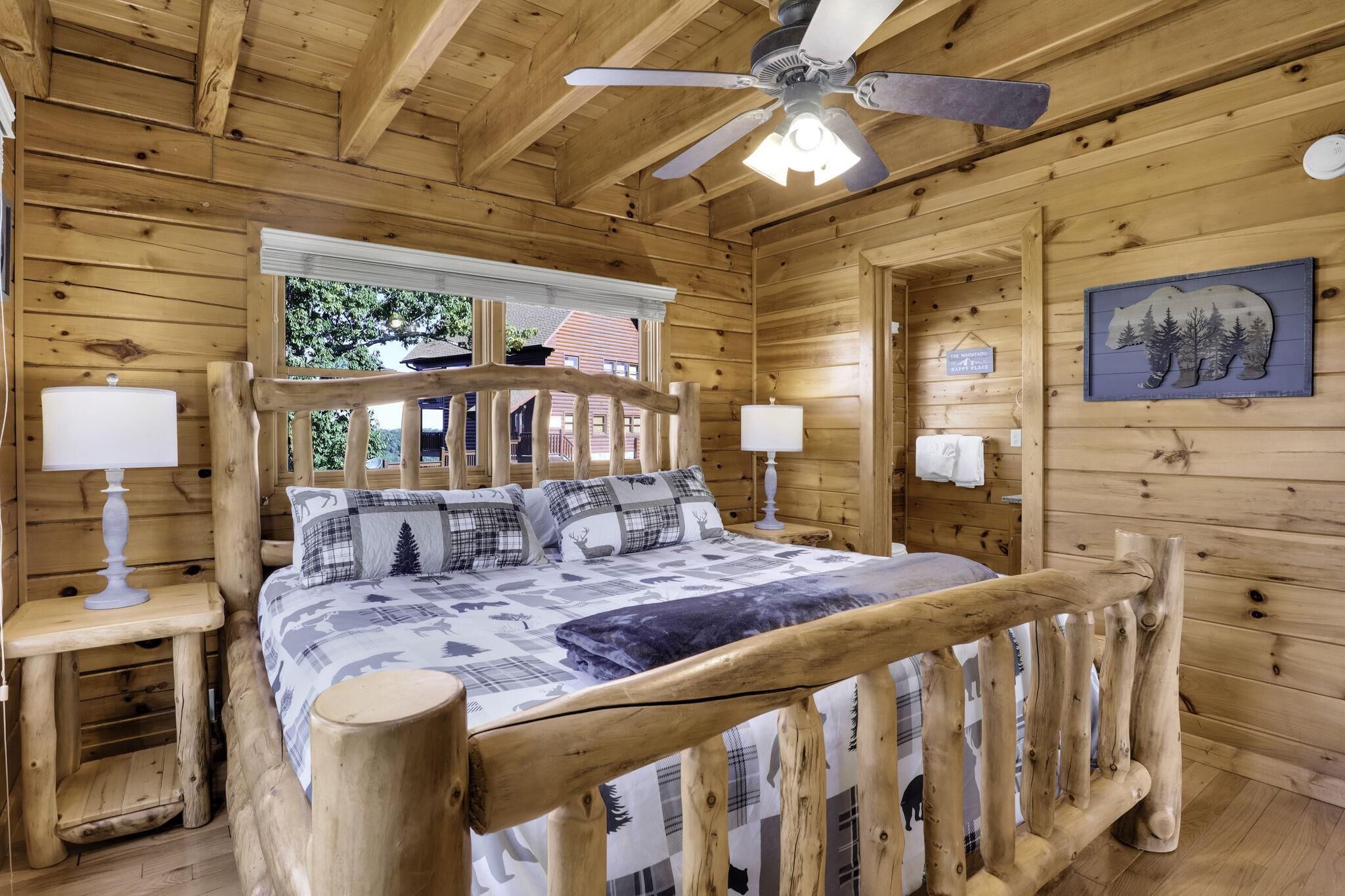 A wooden cabin bedroom with a bed, two bedside tables, a ceiling fan, and a window with a view of the outside.