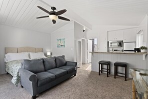 A bedroom with a bed, a gray sofa, a ceiling fan, and a kitchenette with a microwave and a coffee maker.
