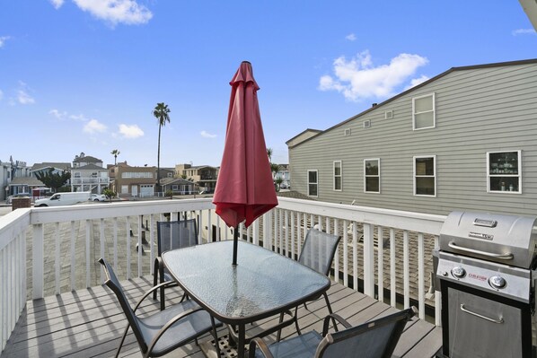A deck with a glass table and chairs, a red umbrella, and a grill.