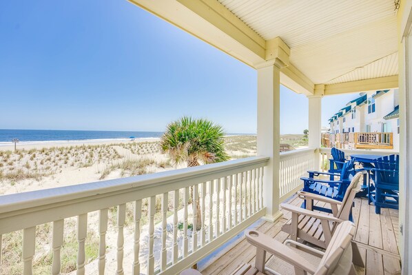Blick auf einen Strand von einer Veranda mit weißen Geländern, blauen Stühlen und einer Palme.