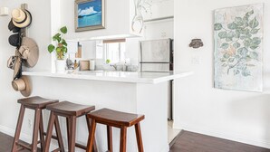 Cocina moderna con encimera blanca, refrigerador de acero inoxidable y una planta colgante. Hay tres taburetes de madera y un perchero en la pared.