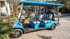 Une voiturette de golf bleue, à deux places et dotée d’un toit, est garée sur une surface pavée, avec un bâtiment et des arbres en arrière-plan.