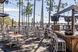 Un espace de restauration en plein air avec des tables et des chaises, un comptoir de bar, et une vue sur l’eau et les bateaux au loin.