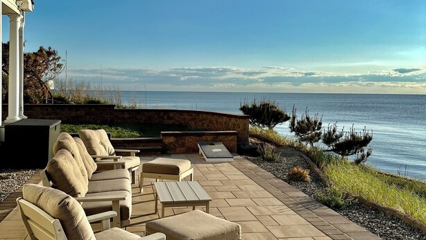 Een terras buiten met beige fauteuils en tafels, een stenen muur en uitzicht op de oceaan.