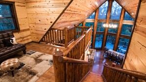 A wooden cabin interior with a loft bedroom, a dining area, and a living room with a leather sofa and ottoman. - Starry Respite: Hot Tub, 3 Fireplaces, Ping Pong, Foosball, Arcade, Pets allowed (Broken Bow)