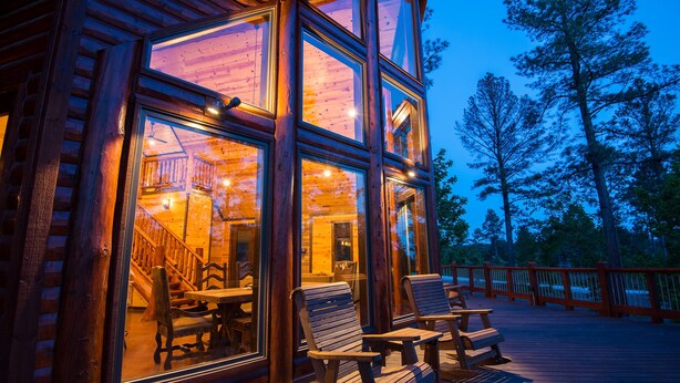 A wooden cabin with large windows, a deck with chairs, and a staircase leading to the interior. - Starry Respite: Hot Tub, 3 Fireplaces, Ping Pong, Foosball, Arcade, Pets allowed (Broken Bow)