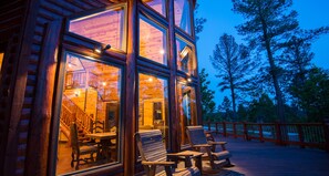 A wooden cabin with large windows, a deck with chairs, and a staircase leading to the interior. - Starry Respite: Hot Tub, 3 Fireplaces, Ping Pong, Foosball, Arcade, Pets allowed (Broken Bow)