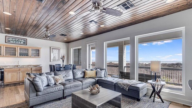 A spacious living area with a large gray sectional sofa, a coffee table, and a view of the outdoors through large windows. - The Beachside - Banyan Beach, Ocean View & Short walk to Beach (Port Aransas)