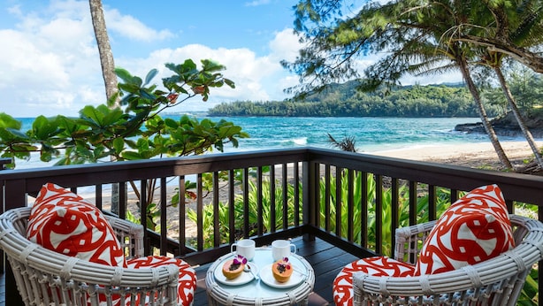 A deck with wicker chairs and a table, overlooking a beach and ocean. - Haena Beach House: Private Luxury Beachfront Home w/ Breathtaking Views (Hanalei)