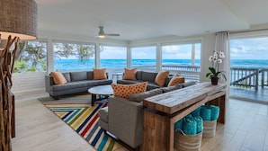 A spacious living area with a large window overlooking the ocean, a gray sofa set, a wooden coffee table, and a colorful striped rug. - Haena Beach House: Private Luxury Beachfront Home w/ Breathtaking Views (Hanalei)
