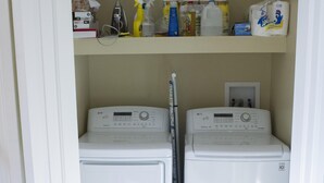 A laundry room with two front-loading washing machines, a dryer, and a shelf above containing cleaning supplies. - Retreat-Like Setting (Trumansburg)