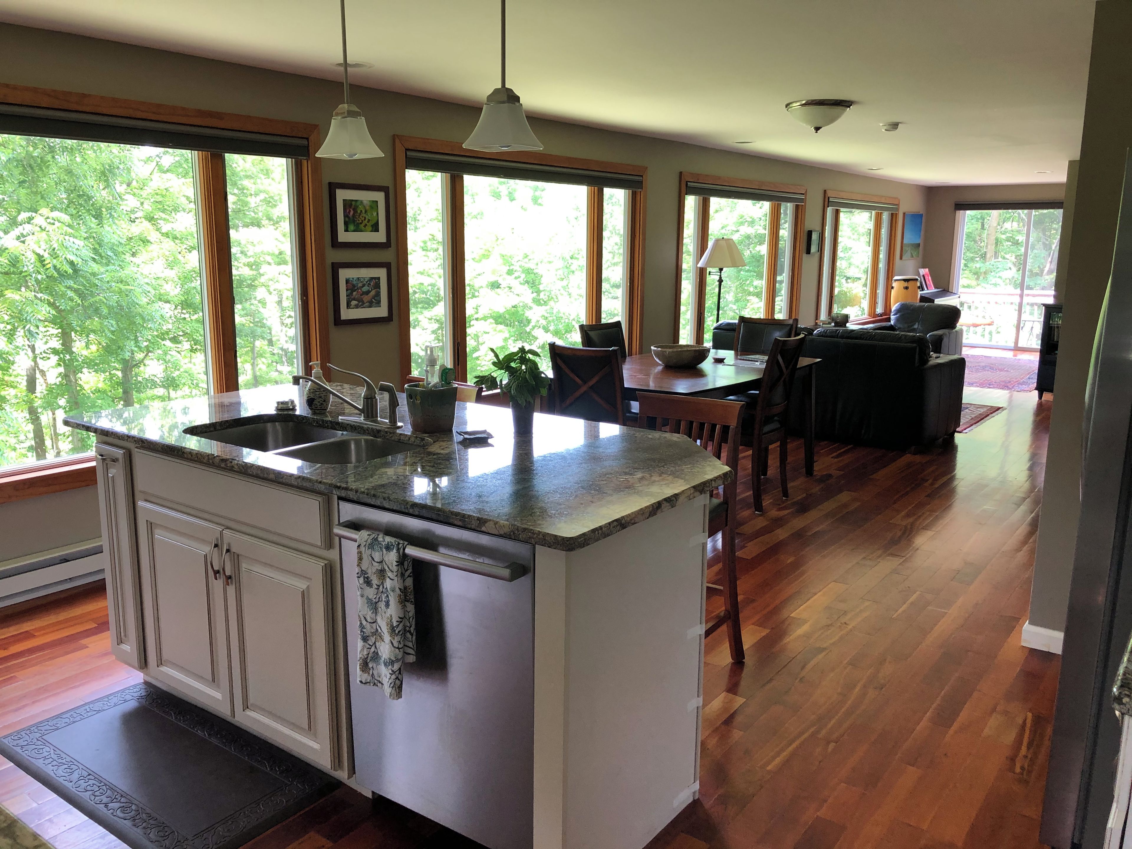 A kitchen with a central island, a dining area with a table and chairs, and large windows with a view of greenery.