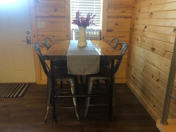 A wooden dining table with four chairs, a vase with flowers, and a window with blinds.