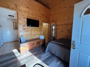 A wooden-paneled room with a bed, a dresser, a television, and a door. - Frenchy's Hideaways Cabin #5 (Custer)