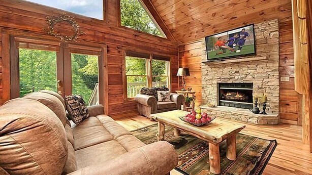 Interior acogedor de una cabaña de madera con chimenea de piedra, televisor grande montado en la pared y un sofá de cuero.