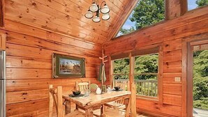 Interior de una cabaña de madera con una mesa de comedor, sillas, un refrigerador y un amplio ventanal con vistas a follaje.