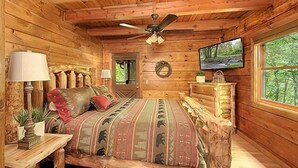 Un dormitorio tipo cabaña de madera con una cama, un televisor, un ventilador de techo y una ventana con vistas a árboles.