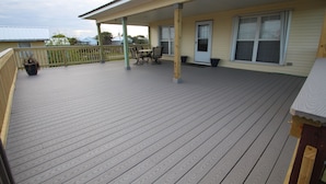 Une terrasse couverte avec un plancher en bois, un coin salon avec des chaises et une table, et une porte située sur la droite.