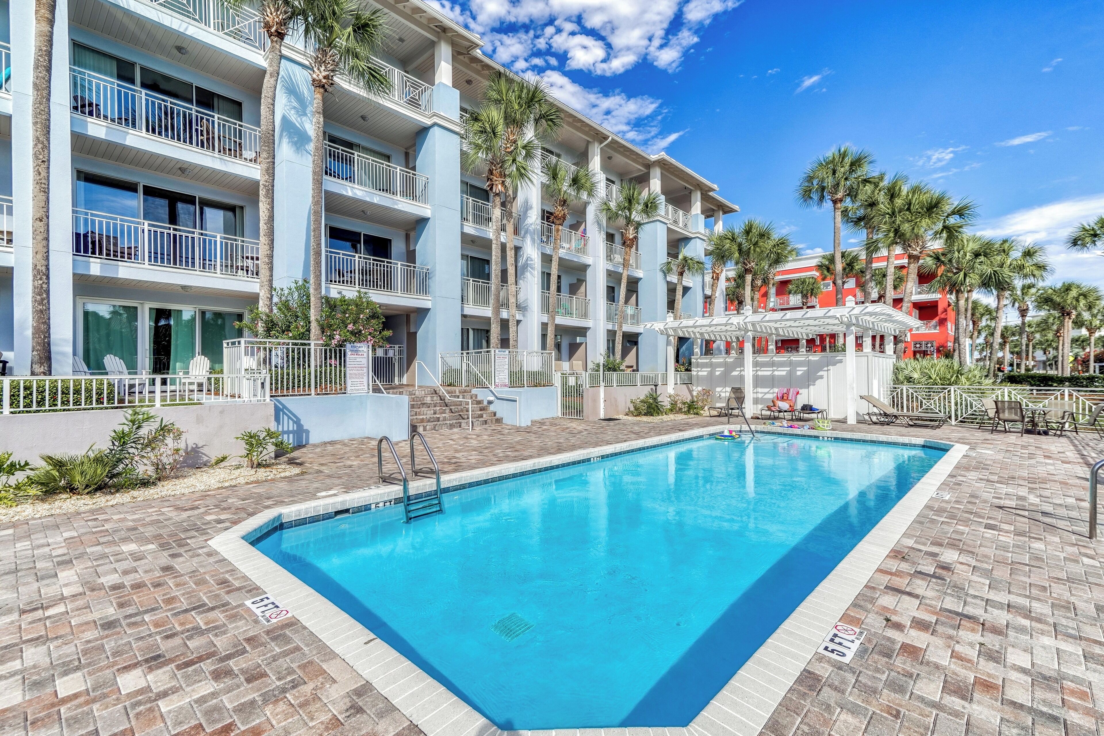 A hotel with a swimming pool, multiple balconies, palm trees, and a red building in the background.