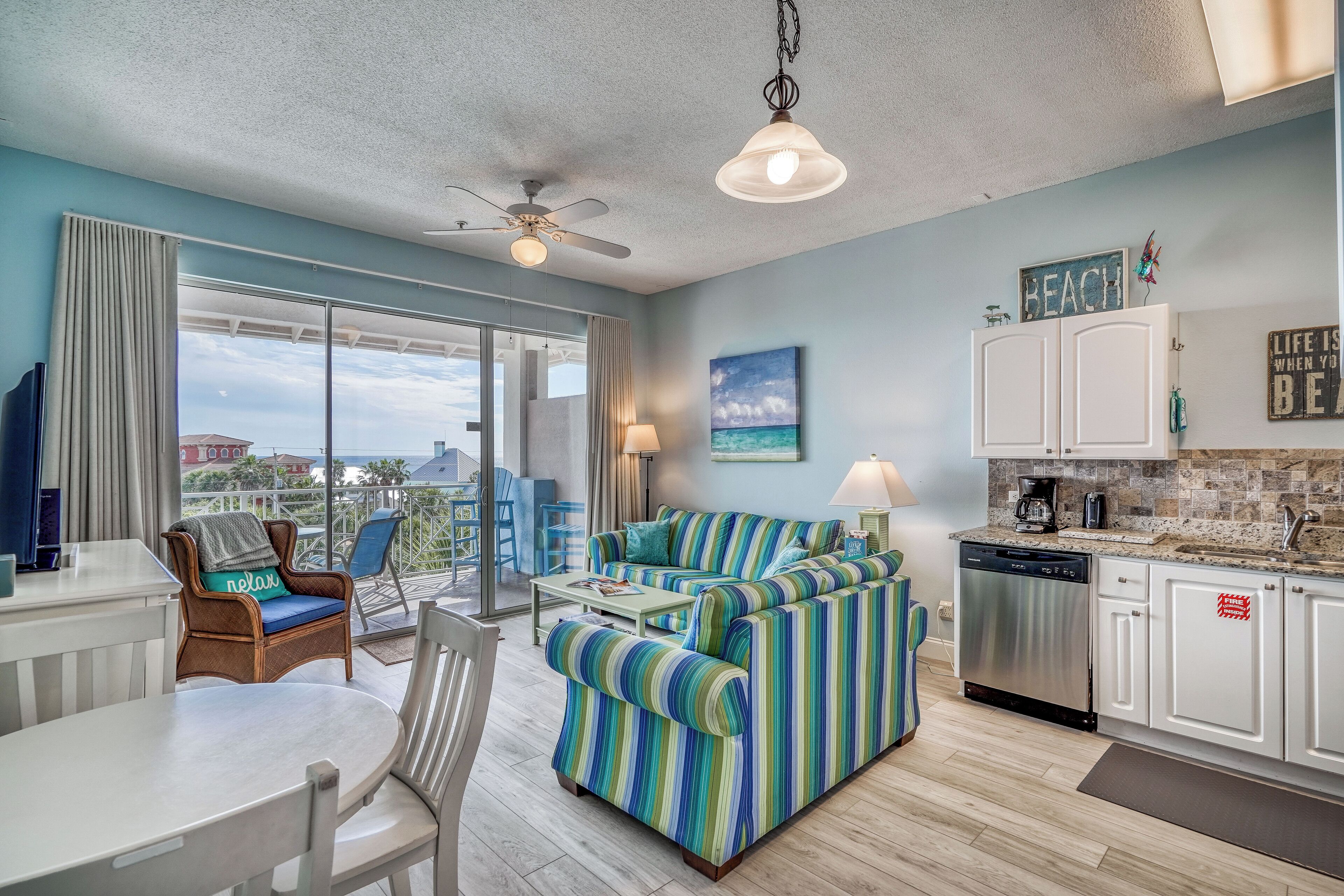 A modern living room with a striped sofa, a dining table, and a kitchen area with white cabinets and a dishwasher.